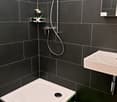 modern style bathroom with black tiled walls, white floor, walk-in shower, vanity with sink and a shower head, towel rack, floor mat