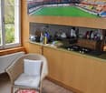 A compact kitchen with a small dining area, wooden cabinets, and a large window providing natural light. The room includes a wicker chair, a table with a white top, a beige carpet, and a radiator for heating. A large photo or painting of a stadium is moun