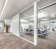 The image shows a modern, open-plan office space with a reception area and a conference room visible through glass walls. The office has a clean, minimalist design with gray carpeting, white walls, and large windows that let in natural light. The receptio