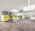 Bright and open office space with large windows, skylights, and a modern, minimalist design. The space features a yellow sofa, potted plants, and a foosball table, creating a relaxed and collaborative atmosphere. The flooring is a patterned carpet tile, a