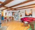 Fireplace, wooden flooring, wooden beams, red couch, dining table with chairs, bookshelf, paintings, wooden coffee table