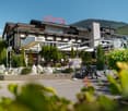 A large hotel building with a prominent 'HOTEL' sign on the roof, surrounded by lush greenery and mountains in the background. The hotel has a terrace and outdoor seating area, and there is a bicycle rack in the foreground.