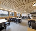Large dining room with wooden ceiling and walls, large windows overlooking a scenic mountain landscape, multiple dining tables and chairs, kitchen area visible in the background