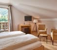 bedroom with two single beds, desk with lamp and tv, 2 wicker armchairs, small table, balcony with chairs and table, floor lamp, wooded floor