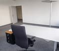 The image shows an empty office space with a desk, an office chair, and a floor lamp. The room has a gray carpet floor and a suspended ceiling with a grid pattern. The walls are white, and there is a door visible in the background.
