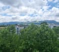 The image shows a panoramic view of a city surrounded by lush greenery and rolling hills. The cityscape is dominated by modern multi-story buildings and apartments, with a mix of residential and commercial structures. The sky is partly cloudy, creating a