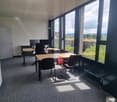 Large open office space with floor-to-ceiling windows providing a scenic view of the surrounding landscape. The room is furnished with multiple desks, chairs, and computer equipment, suggesting it is used as a workspace or office. The overall design appea