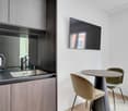 Modern kitchen with dark wood cabinets, stainless steel appliances including a dishwasher, and a small dining area with a round table and velvet chairs.