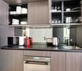 Modern kitchen with gray wood cabinets, black countertop, built-in appliances including a dishwasher and coffee maker, and open shelving displaying dishes and glassware.