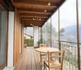 Wooden floor, glass walls, balcony railing, wooden table and chairs, outdoor furniture, potted plant, wooden ceiling, view of mountains