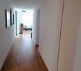 The image shows a long, well-lit hallway with white walls and a hardwood floor. There are several doors along the hallway, and at the end of the hallway, there is a room with a table and chairs, suggesting this is an office or workspace area.