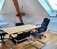 Attic office space with wooden beams, large windows, wooden table, black office chairs, and a small plant on the table.