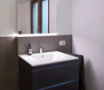 Bathroom: white tiled floor, white sink, large mirror, dark grey cabinetry, built-in towel rack, power outlet, wooden window.