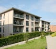 Modern 3-story apartment complex, white exterior, glass balconies, hedged garden, paved walkway, green lawn, plants, and shrubs.