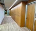 The image shows a long hallway with wooden paneled walls and doors on both sides. The floor is covered with a light-colored laminate or vinyl flooring. The hallway is well-lit with recessed lighting fixtures in the ceiling.