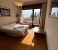 A bedroom with a bed, a glass table, a lamp, and large windows leading to a balcony with a view of the surroundings.