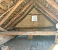 The image shows the interior of an attic space. The ceiling is made of exposed wooden beams and rafters, creating a rustic and traditional appearance. The floor is covered with wooden planks, and there is a small window that allows natural light to enter