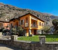 Two-story orange house with balconies, surrounded by stone walls and landscaping, set against a backdrop of mountains