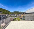 The image shows a balcony or terrace overlooking a scenic mountain town. The balcony has a tiled floor and metal railings. In the distance, there are colorful buildings and lush green trees against a backdrop of mountains and a clear blue sky.