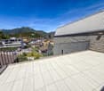 The image shows a balcony or terrace with a view of a town surrounded by mountains. The balcony has a tiled floor and a metal railing. The building has a corrugated metal roof and appears to be part of a larger residential or commercial complex.