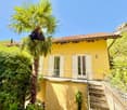 2 story yellow house, red roof, balcony, windows with white shutters, steps leading to entrance, surrounded by trees and plants