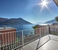 Outdoor balcony with railings, overlooking a lake, mountains, and a clear sky. There is a palm tree and a building in the background.
