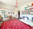 Large red rug, white walls, two blue couches, coffee table, hanging lamps, dining table with chairs, shelves with books and decorative items, plants in pots, mirrors, radiator, open door leading to another room