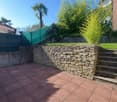 Stone retaining wall, brick paved patio, plants, stone steps, fence, view of trees