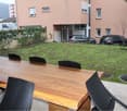 A garden with a wooden table and black chairs, a grassy area, and a building with parked cars in front.