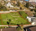 Aerial view of a plot of land with a house on the right side, surrounded by other houses and greenery, with a mountain in the background.
