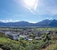 The image shows a panoramic view of a town or city nestled in a valley surrounded by mountains. The landscape features a mix of residential and commercial buildings, with green fields and trees in the foreground. The sky is clear and bright, with some clo