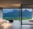 Spacious living room with large floor-to-ceiling windows offering a stunning view of the mountainous landscape. The room features a comfortable sofa, a coffee table, and an additional seating area with a chair. The interior design is minimalist and modern