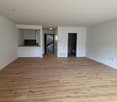Empty room with wooden floor, white walls, kitchen area with cabinets, open door leading to staircase, another door leading to bathroom, power outlets on the wall.