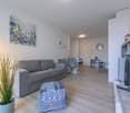 Light colored room, wooden floor, gray couch, ottoman, table with remote control, white walls, large abstract art, dining table, chair, plants, shelf with flowers, TV, lamp, table lamp