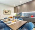 Modern dining room with wooden dining table and blue chairs, wooden cabinets, counter with sink and kitchen appliances, bowl of fruits, wine bottles, wine glasses, and other dining items