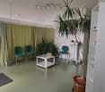 Spacious open-plan living area with green curtains, white walls, tiled flooring, potted plants, and chairs arranged in a seating area.