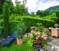 Lush, green garden with tall trees, hedges, and potted plants. Wooden deck with outdoor furniture, including a lounge chair and wicker chairs. Potted flowers and plants on the deck. Scenic mountain view in the background.