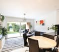 Spacious open-plan living area with large sliding glass doors leading to a balcony or terrace, providing a view of the outdoor greenery. The room features a dining table with chairs, a sofa, and various potted plants. The walls are decorated with artwork,