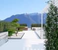 The image shows a well-designed outdoor space with a paved walkway leading to a scenic view of snow-capped mountains in the distance. The space features a bonsai-style tree, lush greenery, and a minimalist design aesthetic. The overall impression is one o