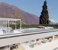 Modern two-story house with a large outdoor terrace and swimming pool. The house has floor-to-ceiling windows offering panoramic views of the surrounding mountains. The exterior features a minimalist white design with a tall pine tree in the foreground.