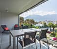 Spacious balcony with outdoor dining area, modern furniture, and panoramic mountain views. The apartment features large windows providing abundant natural light and access to the balcony.
