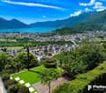 The image shows a panoramic view of a city nestled between mountains and a large lake. The city has a mix of modern high-rise buildings and older structures surrounded by lush greenery. The lake appears to be a vibrant turquoise color, contrasting with th