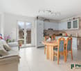 Open-plan living room and kitchen, with large windows and sliding glass doors leading to a balcony. The kitchen has white cabinets and appliances, and the dining area has a wooden table with orange chairs. The living room has a gray sofa and decorative it