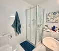 modern bathroom with white tiles, glass shower, white sink, white toilet, blue rug, towel, mirror, toiletries