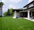 Two-story Mediterranean-style house with a tiled roof, surrounded by a well-manicured lawn and palm trees. The house has a large covered patio or terrace area with outdoor furniture, and the property offers a scenic view of the surrounding landscape.