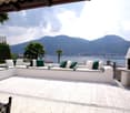 Spacious outdoor terrace with comfortable seating, overlooking a lake surrounded by mountains. The terrace has a tiled floor, a fireplace, and palm trees in the background.