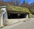 A modern building with a unique, curved roof covered in lush greenery, likely ivy or other climbing plants. The building has a stone exterior and is situated in a wooded, natural setting. There is a paved walkway leading to the entrance of the building, w