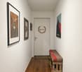 A hallway with white walls, wooden floor, a white door, a bench, and a shoe rack. There are three framed pictures on the wall, one of which has a wreath with the word 'Home' on it.