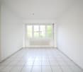 Empty room with white tiles, white walls, large window, white kitchen visible in the background