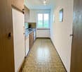 Galley-style kitchen with wooden cabinets, stainless steel appliances including a dishwasher, and a patterned tile floor. The kitchen leads to a hallway with a window providing natural light.
